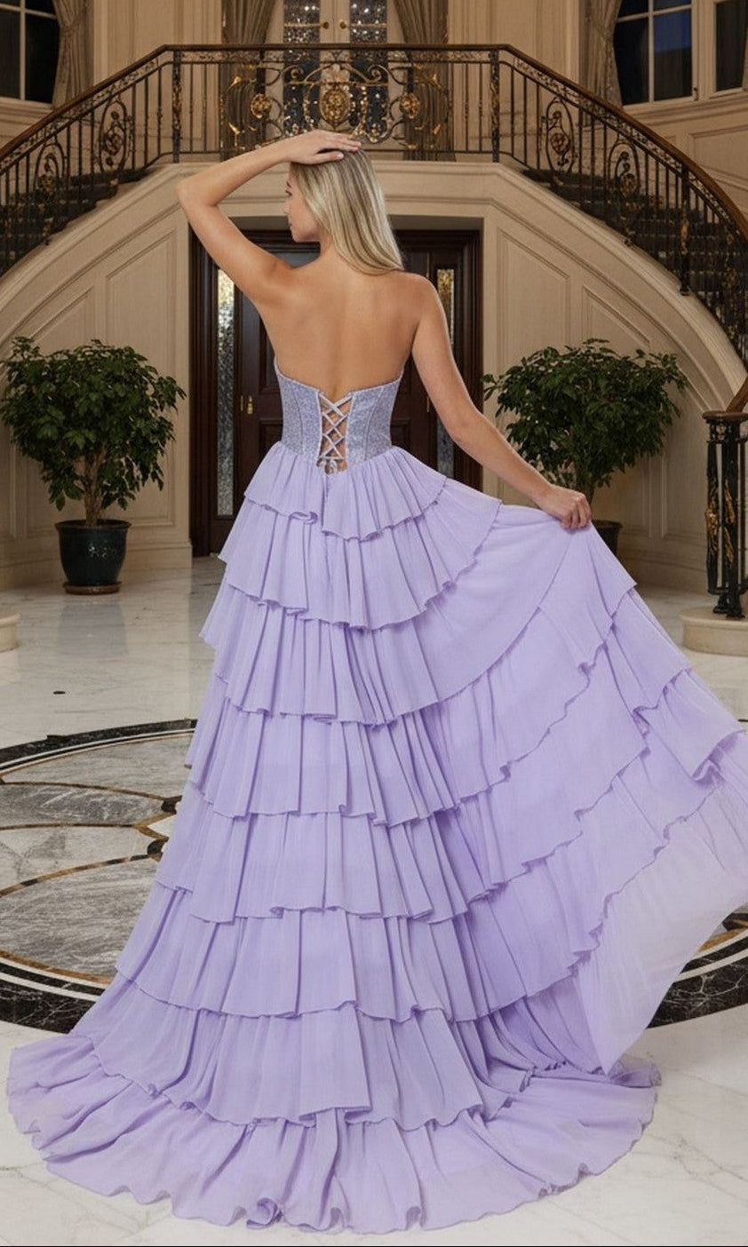 A woman stands with her back to the camera in an ornate foyer, holding the Long Prom Dress: Poly USA 25168—a strapless chiffon gown with a tiered ruffle skirt—on marble floors beside a curved staircase.